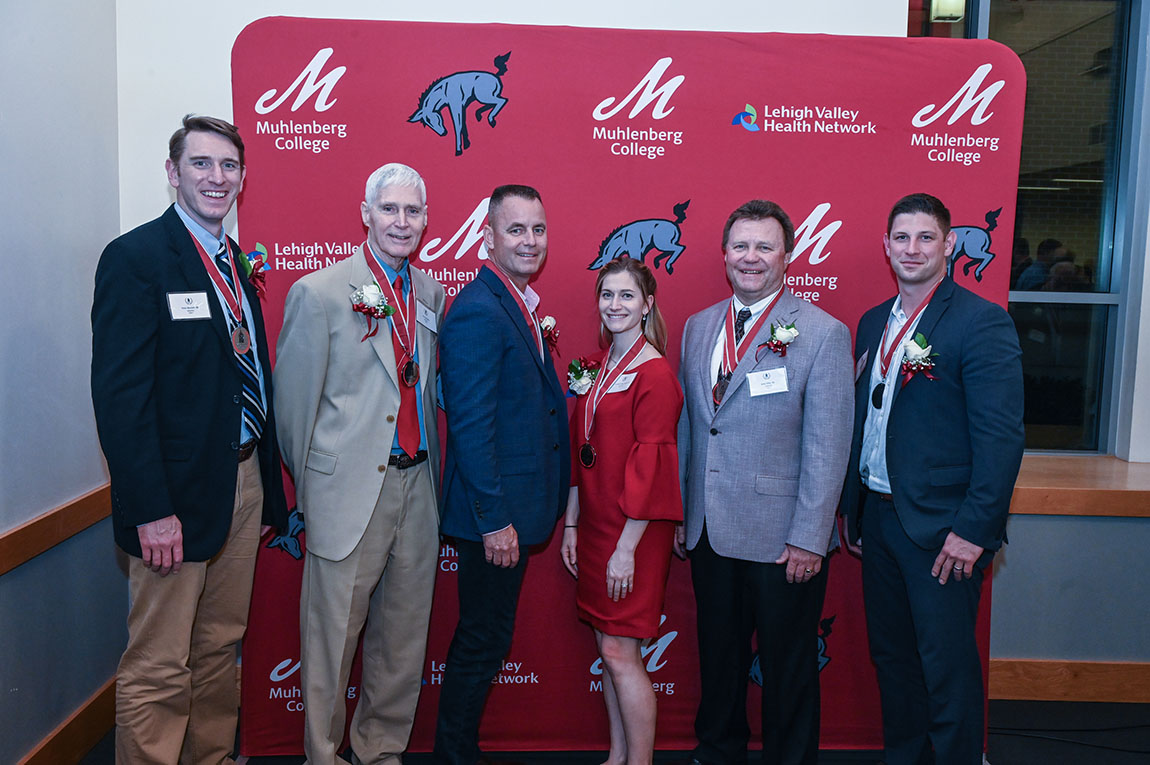 A group of former college athletes smiles in front of a red background